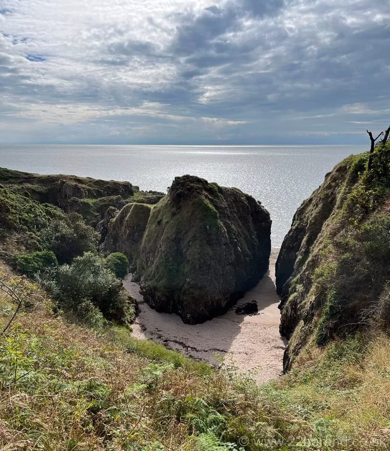 High angle view looking down into a steep, secluded sandy cove nestled between green cliffs on the Solway Coast.