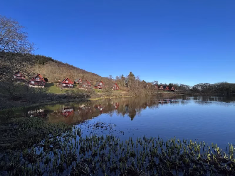 Traditional wooden A-frame holiday lodges perfectly reflected on the still, clear blue water of Barend Loch on a bright day.